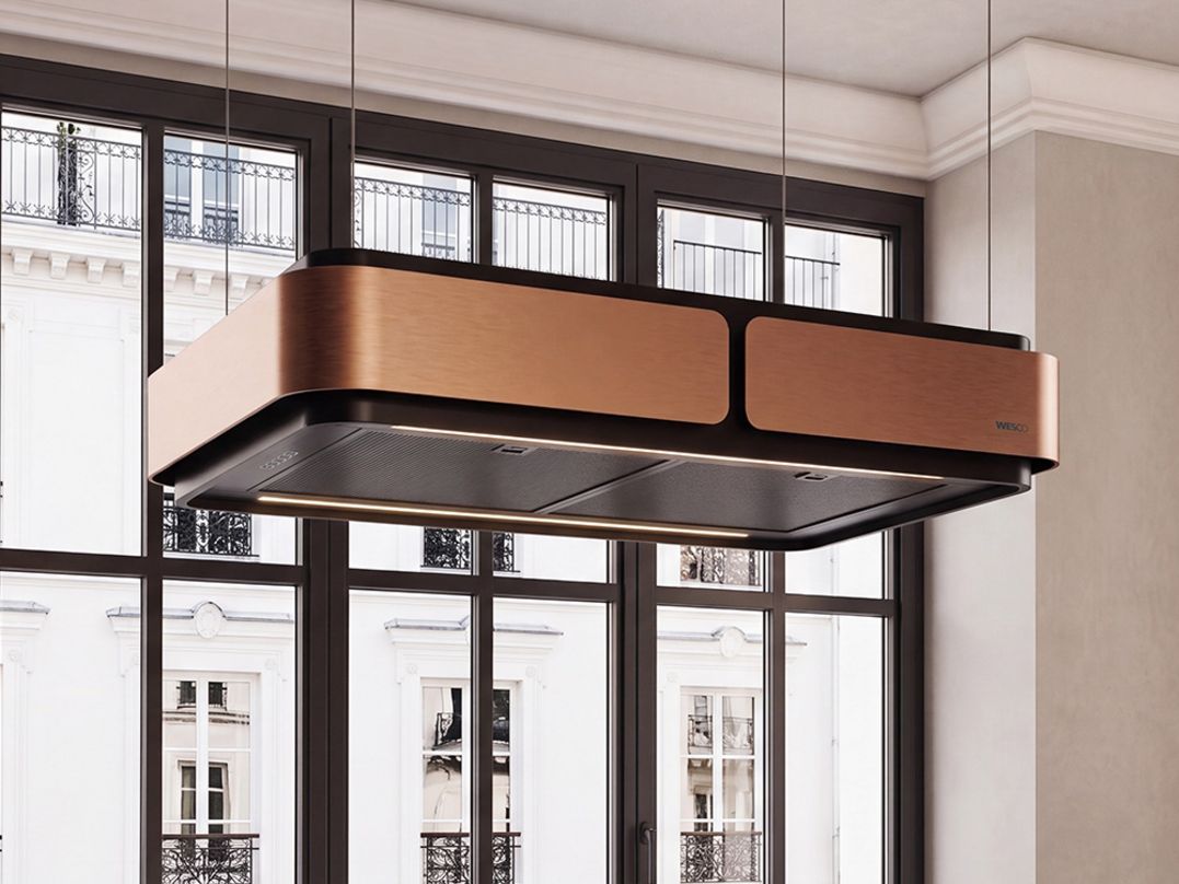 Hotte îlot WESCO Aurea avec finition noir mat et accents cuivrés, flottant élégamment au-dessus d’un îlot de cuisine devant une grande façade vitrée.bandeau lumineux LED intégré.